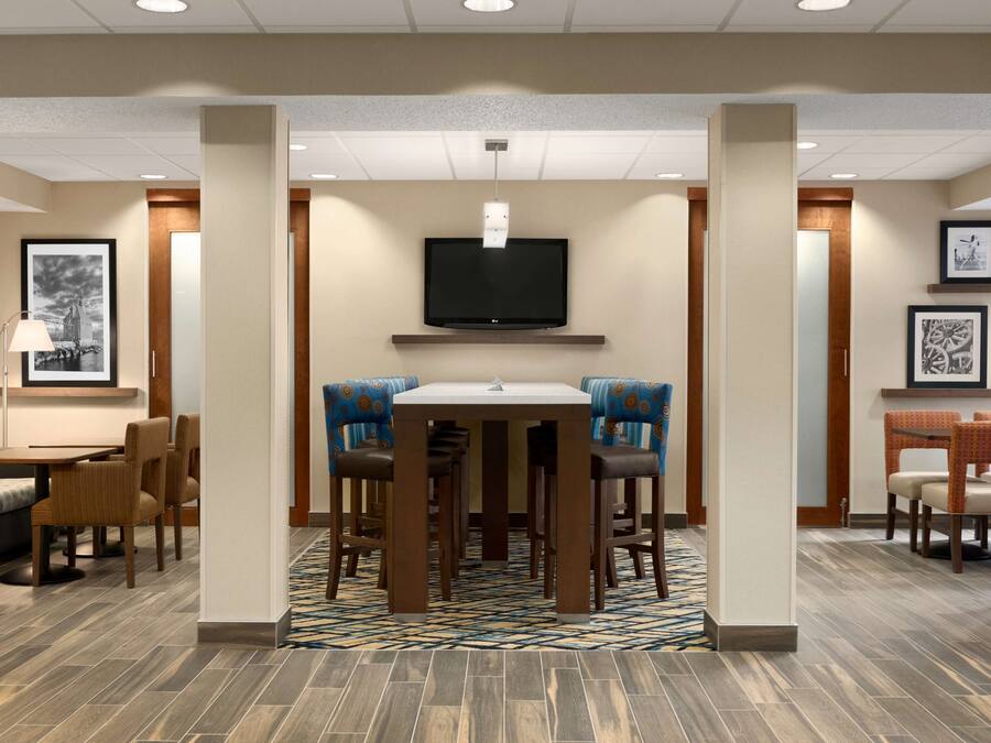 Un vestíbulo de hotel con una mesa de comedor central, taburetes de bar, un televisor de pantalla plana en la pared y cuadros en las paredes.