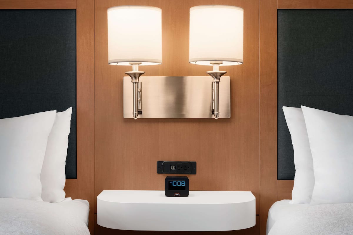A hotel room with a bed, two wall-mounted lamps, a digital clock, and a white bedside table.
