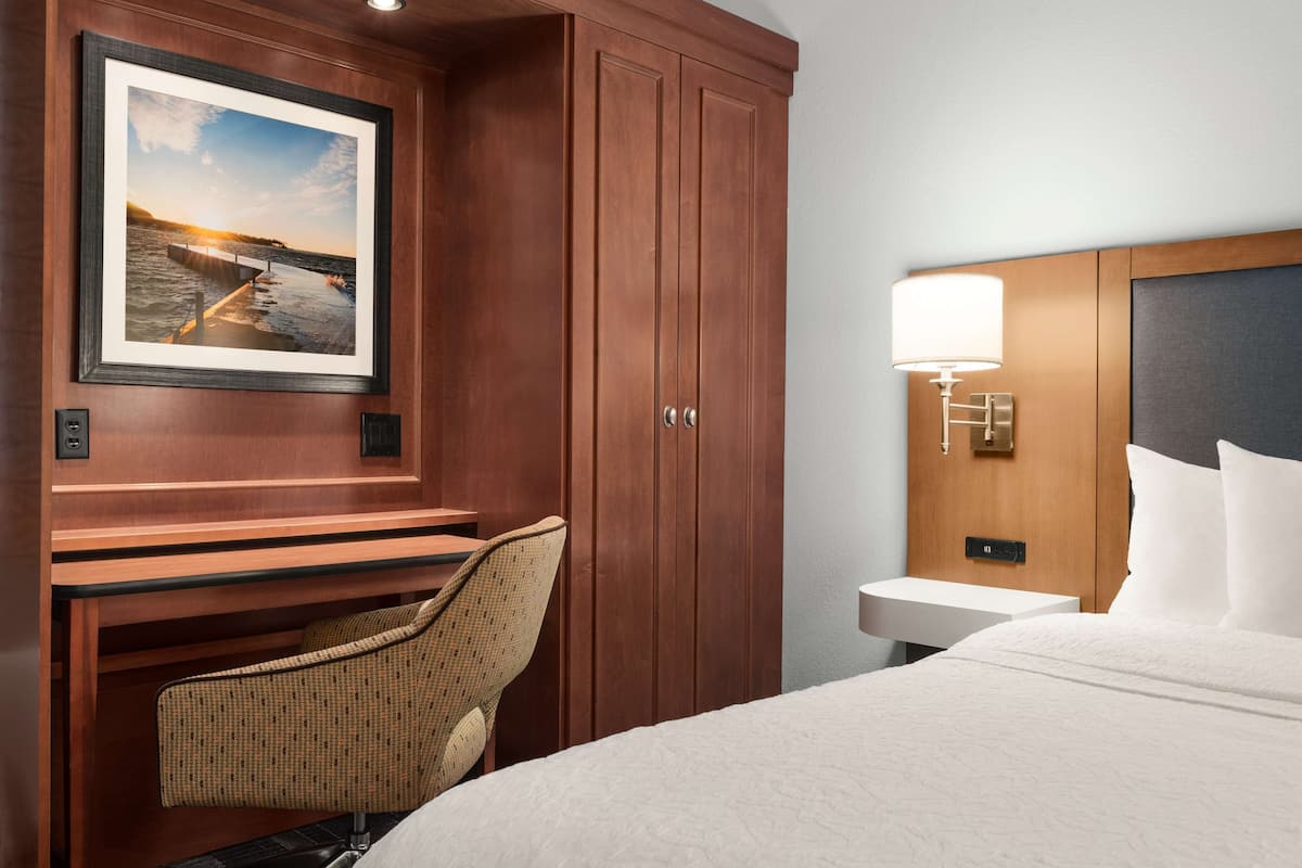 A hotel room with a bed, a desk with a chair, a wooden wardrobe, a framed picture on the wall, a bedside table with a lamp, and a wall-mounted light fixture.