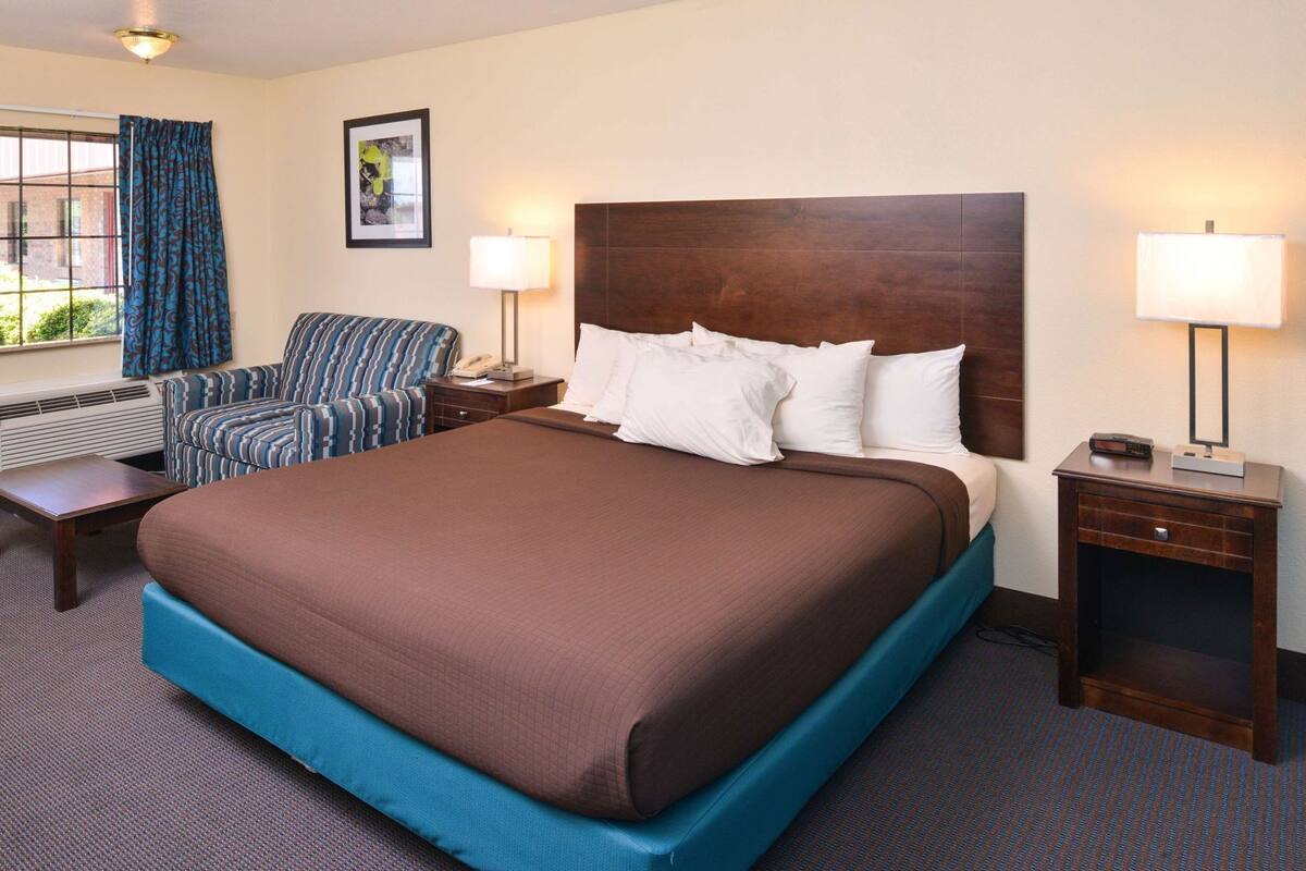 A hotel room with a large bed, a chair, a bedside table, a lamp, and a window with curtains.