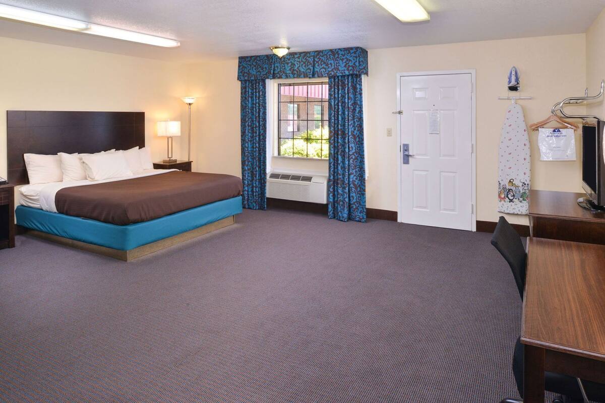 A hotel room with a bed, a desk, a TV, a window with curtains, and a door.