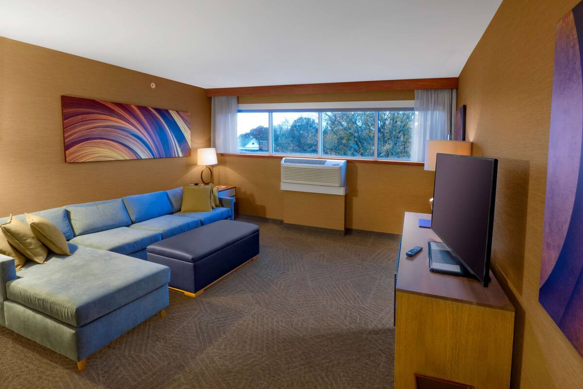 A hotel room with a sectional sofa, a flat-screen TV, a desk, and a large window with a view of trees.