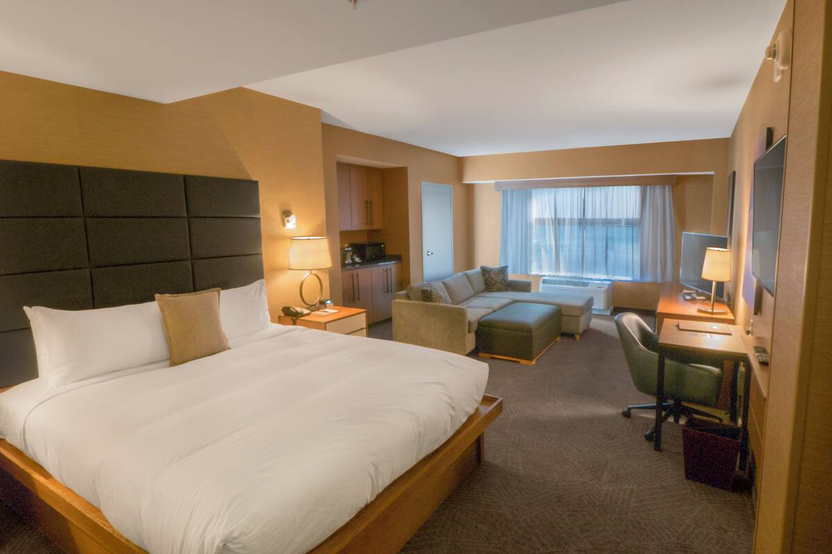 A hotel room with a large bed, a sofa, a desk, and a chair. There is a kitchenette in the corner and a window with curtains.