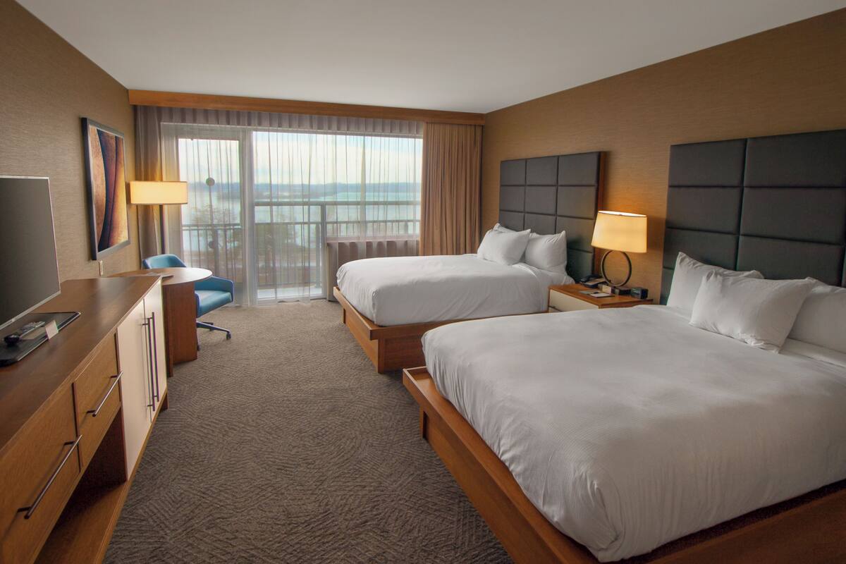 A hotel room with two beds, a desk with a computer, a chair, and a view of the outdoors through large windows.
