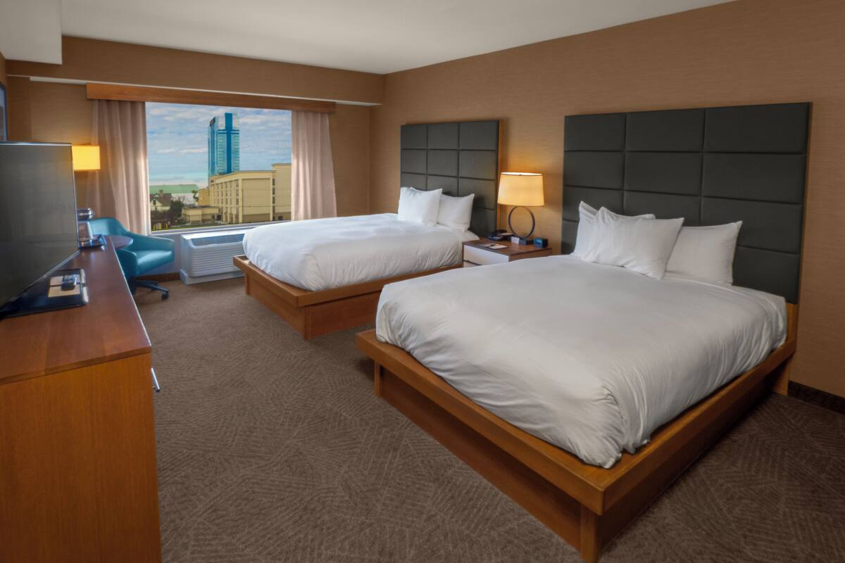 A hotel room with two beds, a desk, a TV, and a window with a city view.
