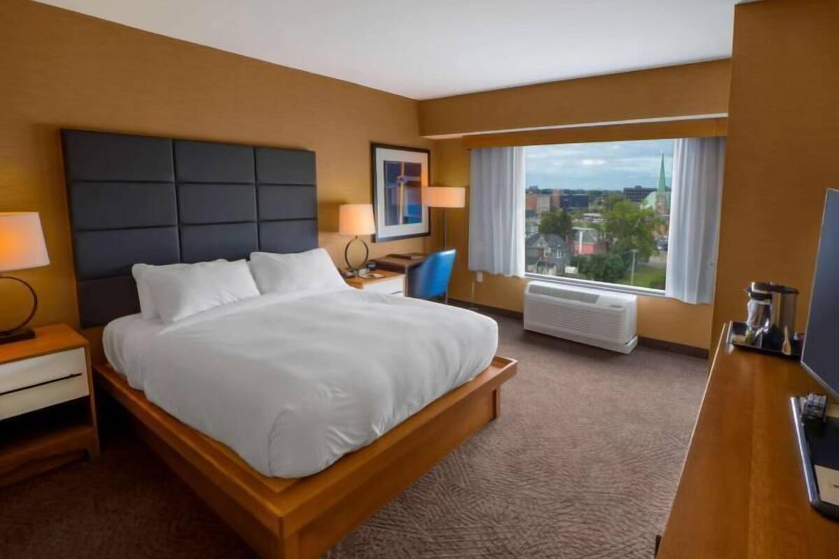 A hotel room with a bed, bedside table, lamp, chair, window, and air conditioning unit.