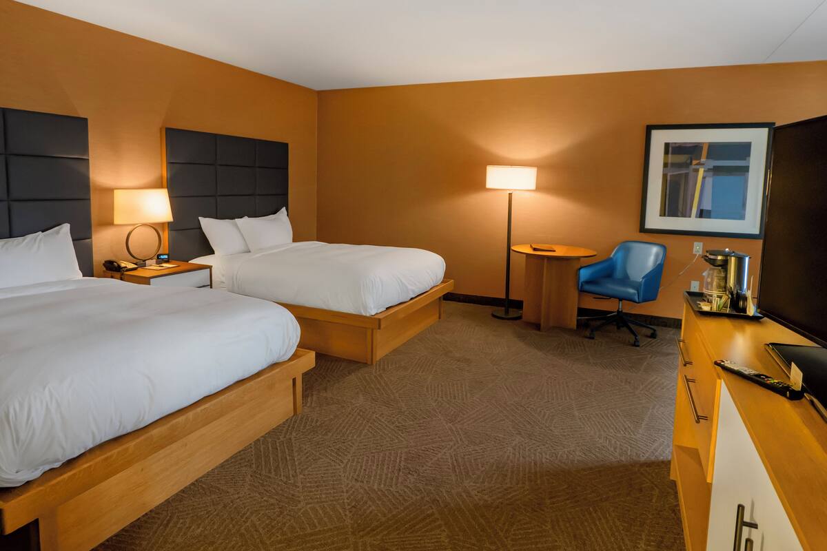 A hotel room with two beds, a desk, a chair, a lamp, and a television.