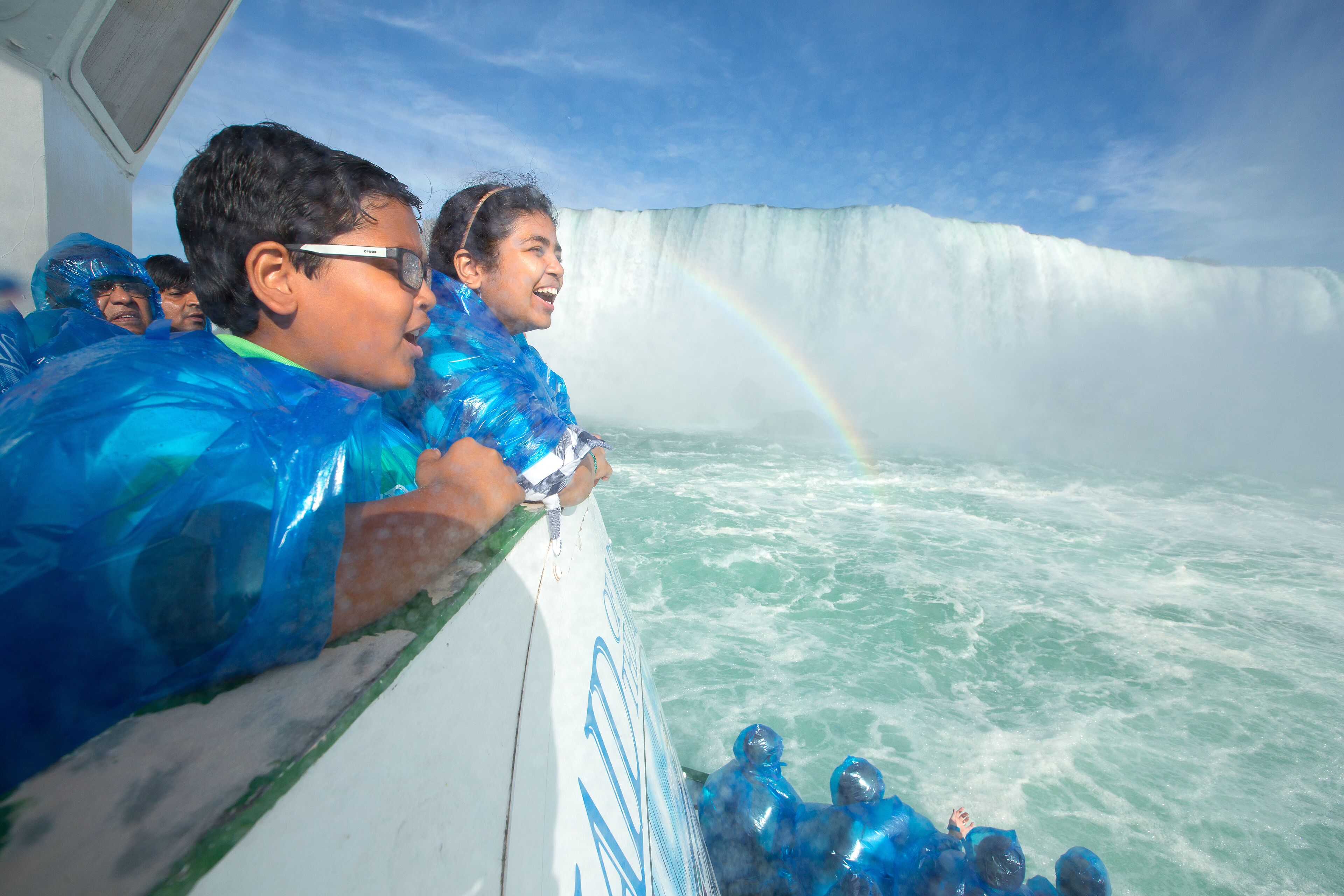 A boat with passengers wearing blue rain ponchos, a large waterfall, and a rainbow in the background.