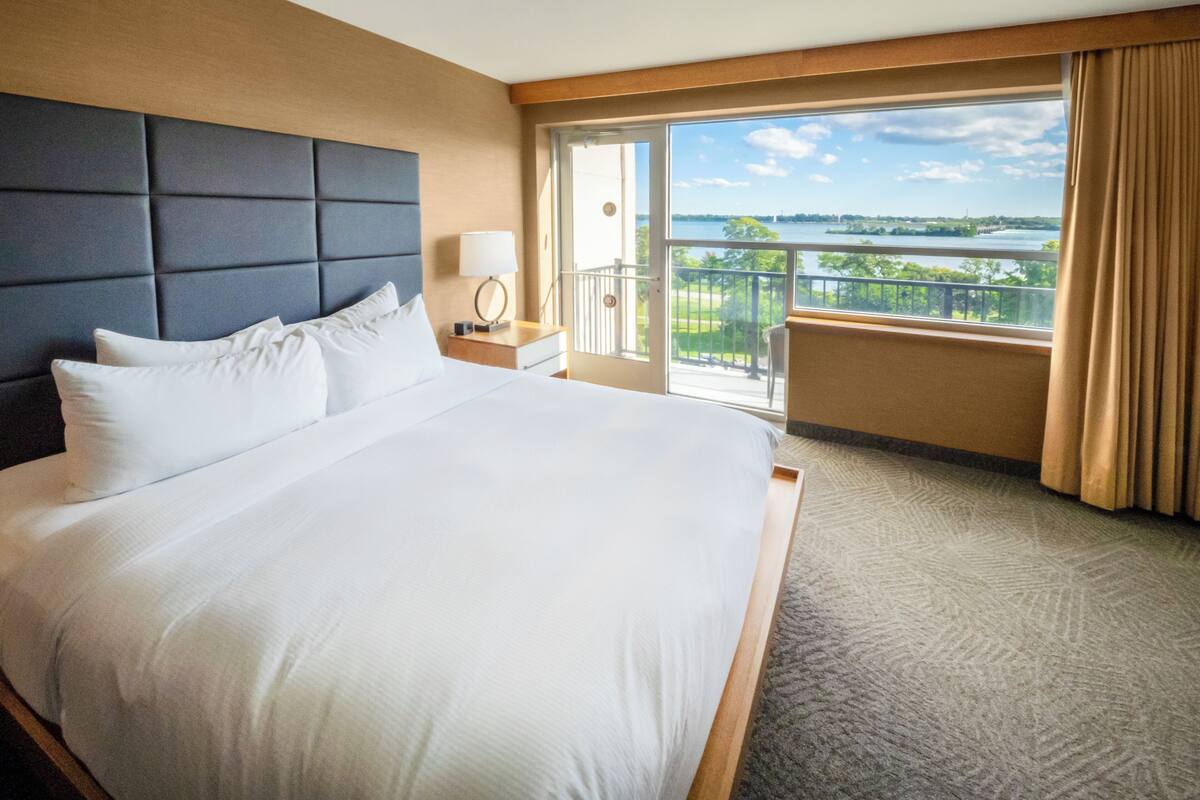 A hotel room with a large bed, a nightstand, a lamp, and a view of a body of water through a balcony.