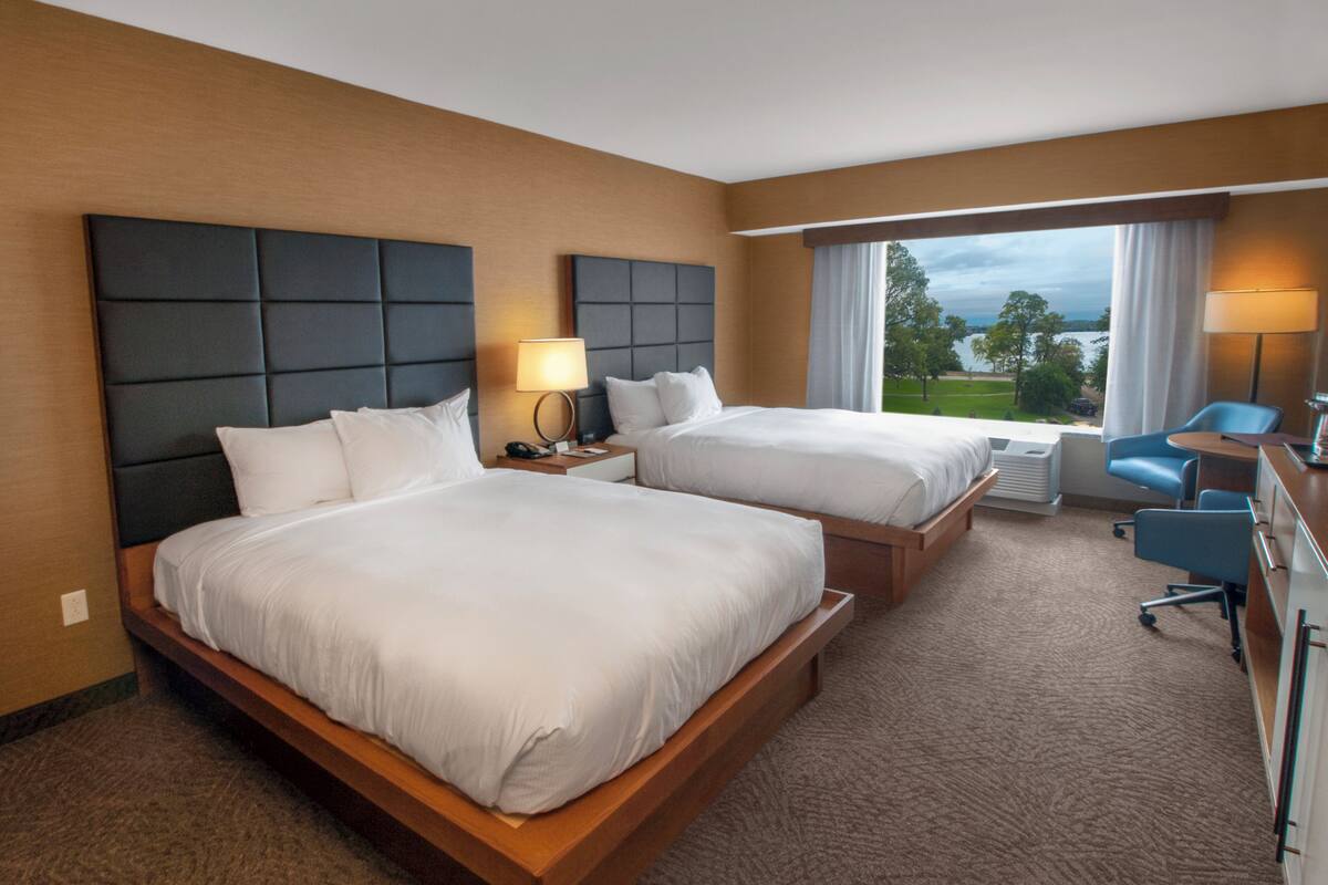 A hotel room with two beds, a desk, a chair, and a view of the outdoors.