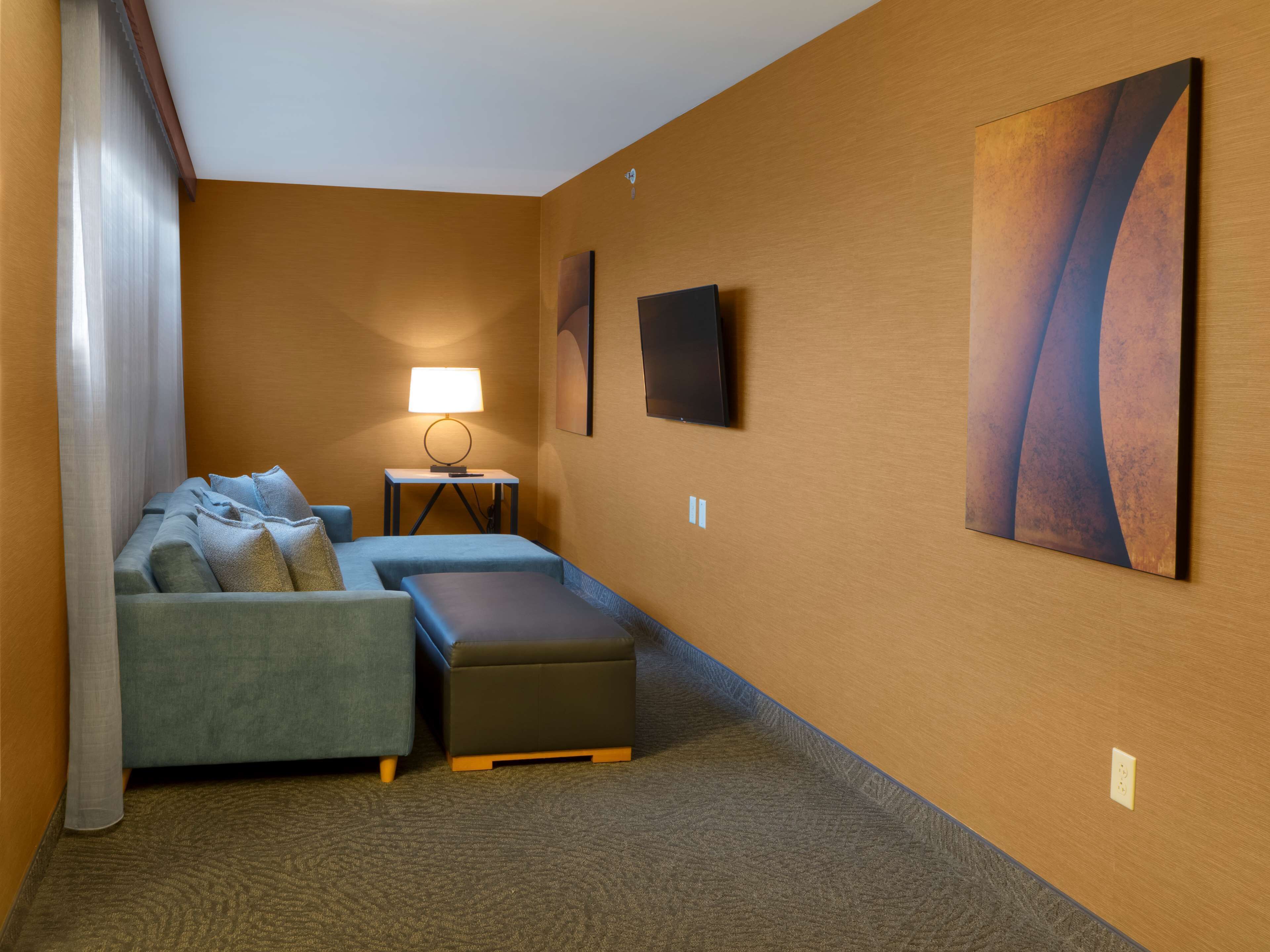 A hotel room with a sofa, ottoman, lamp, and artwork on the wall.