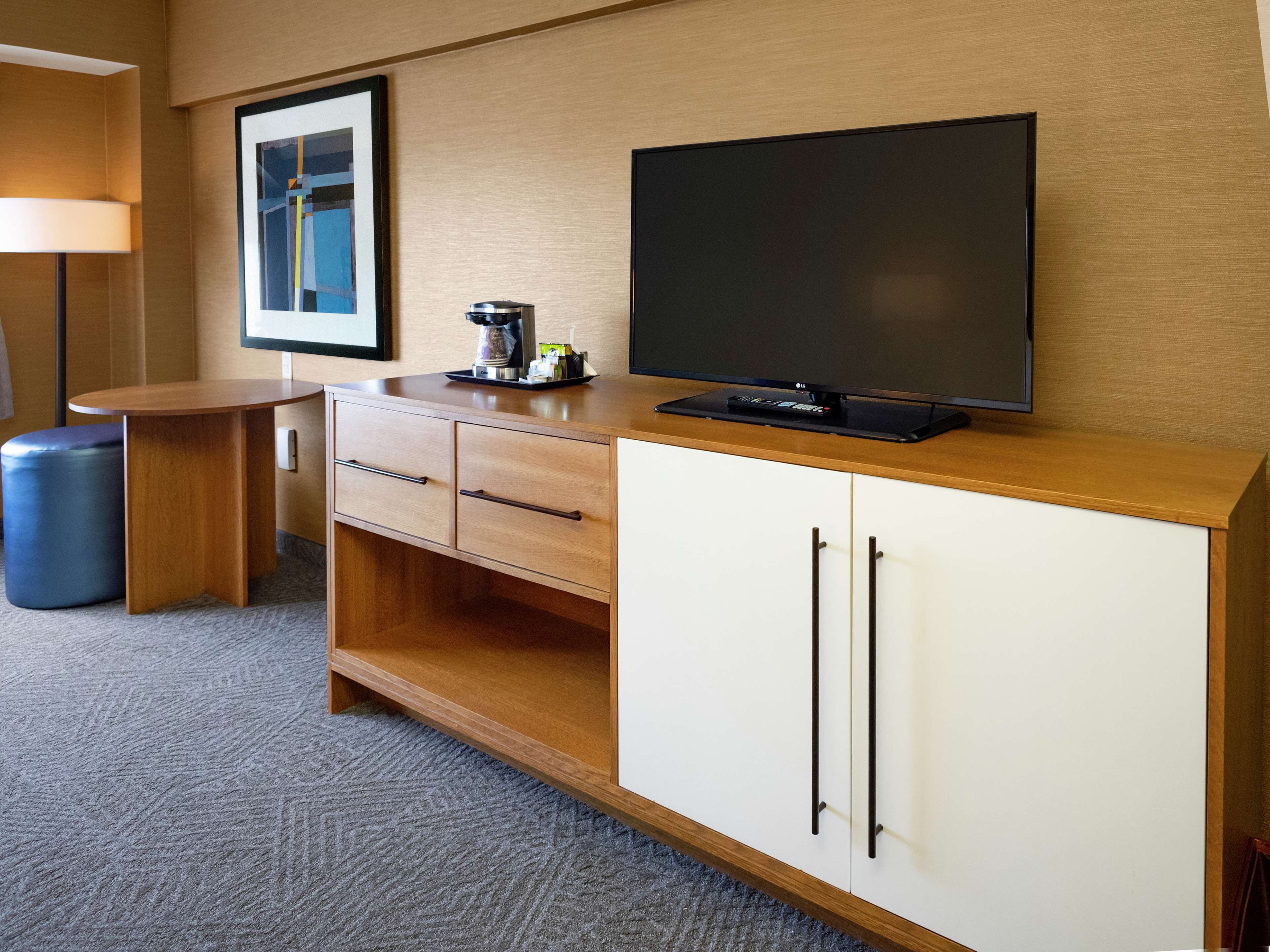 Quarto de hotel com escrivaninha de madeira, televisão de tela plana, cafeteira e quadro pendurado na parede.