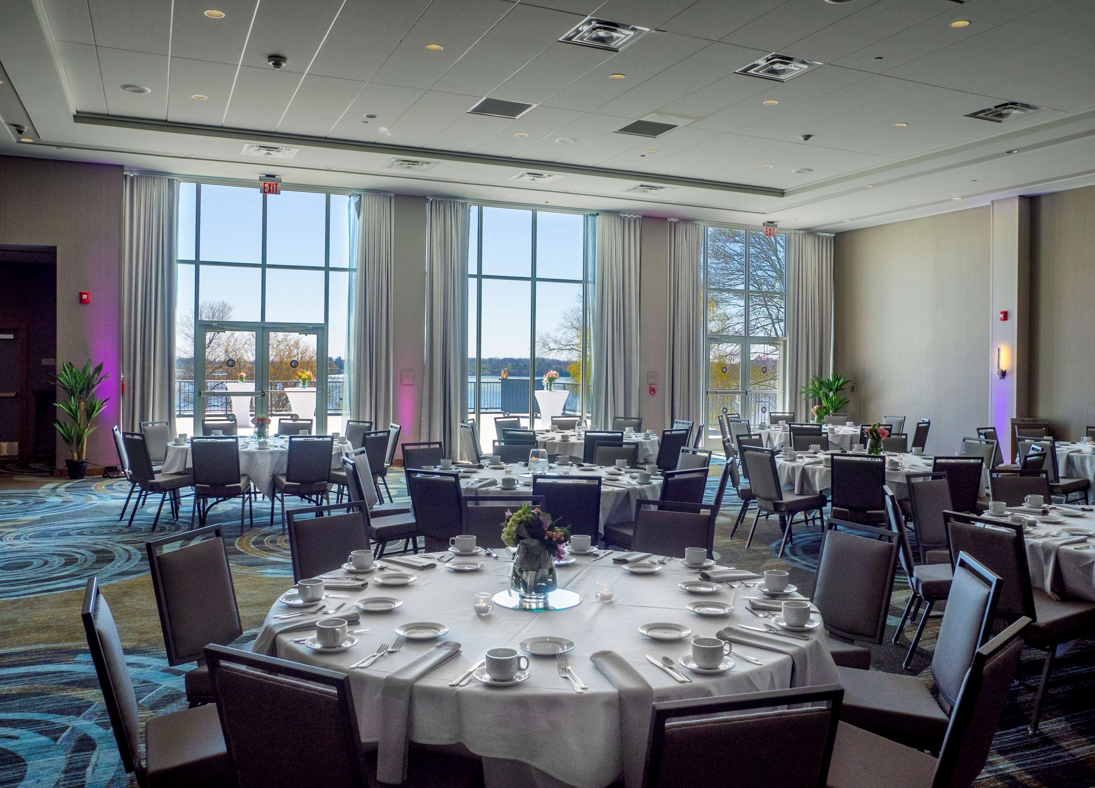 A large conference room with multiple round tables, each set with plates, cups, and cutlery. The room has large windows with curtains, allowing natural light and views of the outdoors.