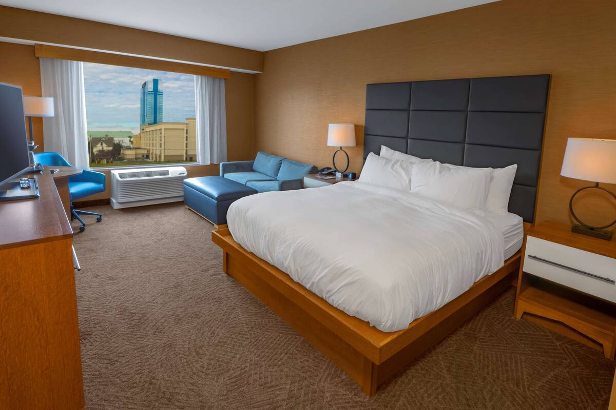 A hotel room with a large bed, a desk with a computer, a chair, a sofa, and a view of a cityscape.