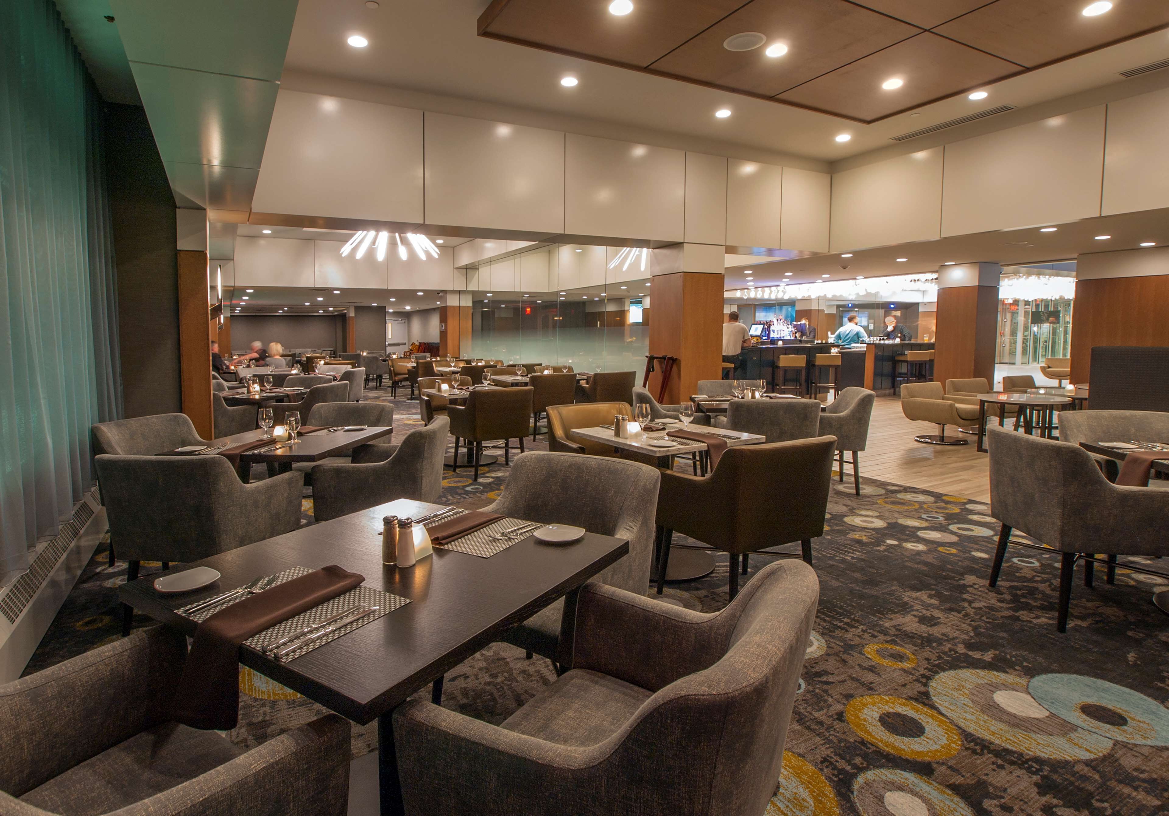 A spacious hotel dining area with multiple tables and chairs, a bar in the background, and a modern ceiling design.