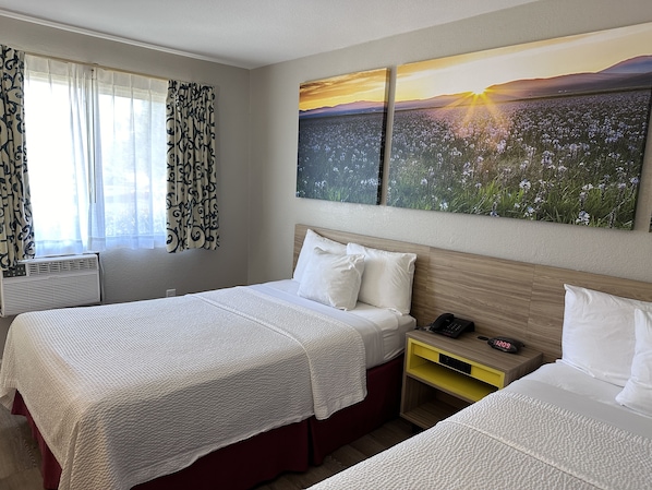 Habitació d'hotel amb dos llits, una finestra amb cortines estampades, un aparell d'aire condicionat, una taula de nit amb un telèfon i un quadre amb un capvespre sobre un camp.
