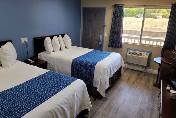 A hotel room with two beds, a window with a view of the outside, a door, and a small table with a chair.