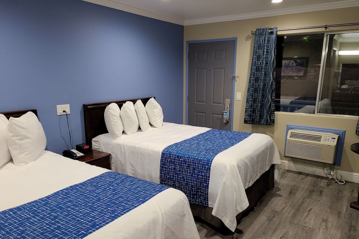 Une chambre d’hôtel avec deux lits, une porte, une fenêtre avec des rideaux, un climatiseur fixé au mur et une table de chevet avec un téléphone.