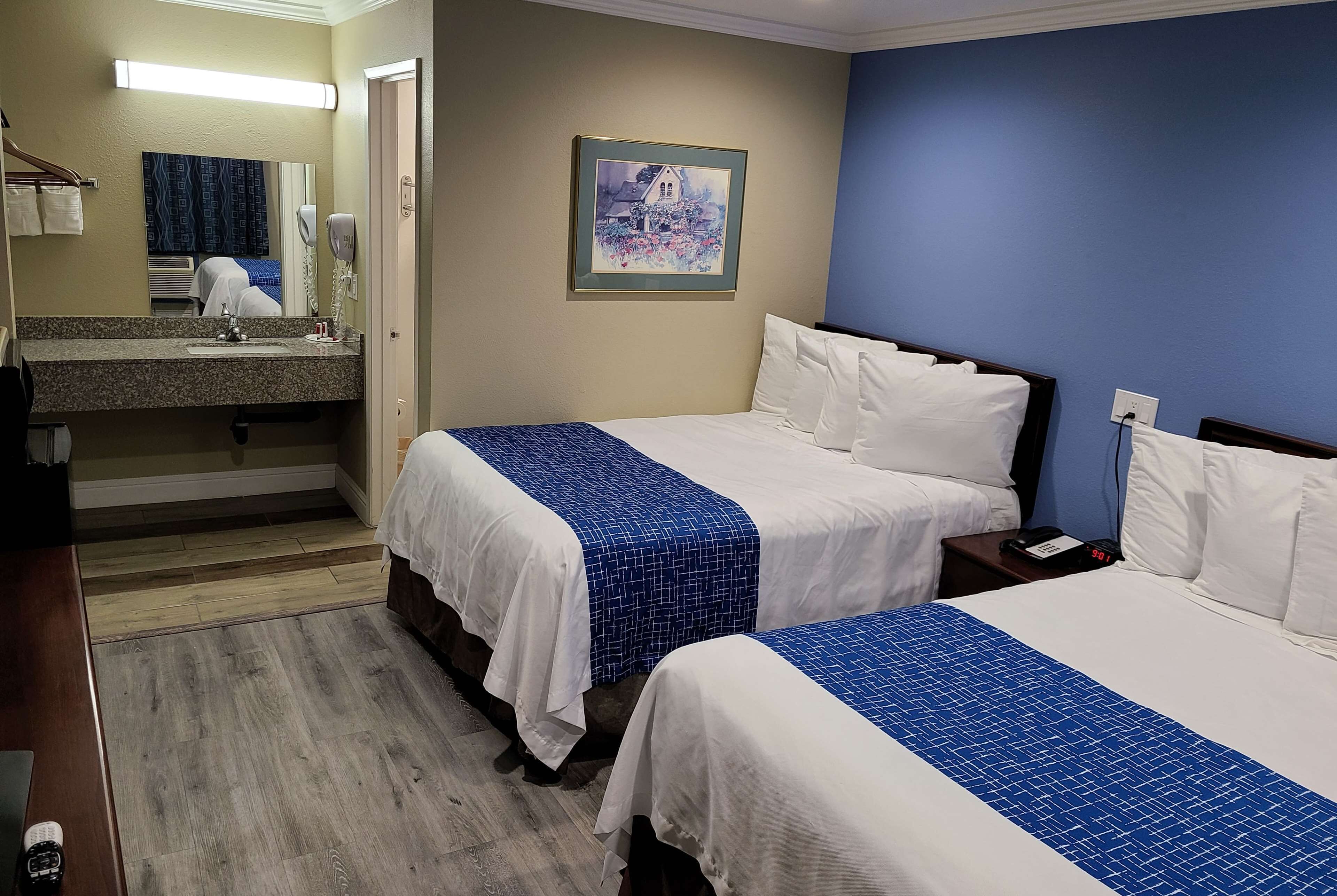 Habitación de hotel con dos camas, un baño con lavamanos y espejo, y un cuadro en la pared.