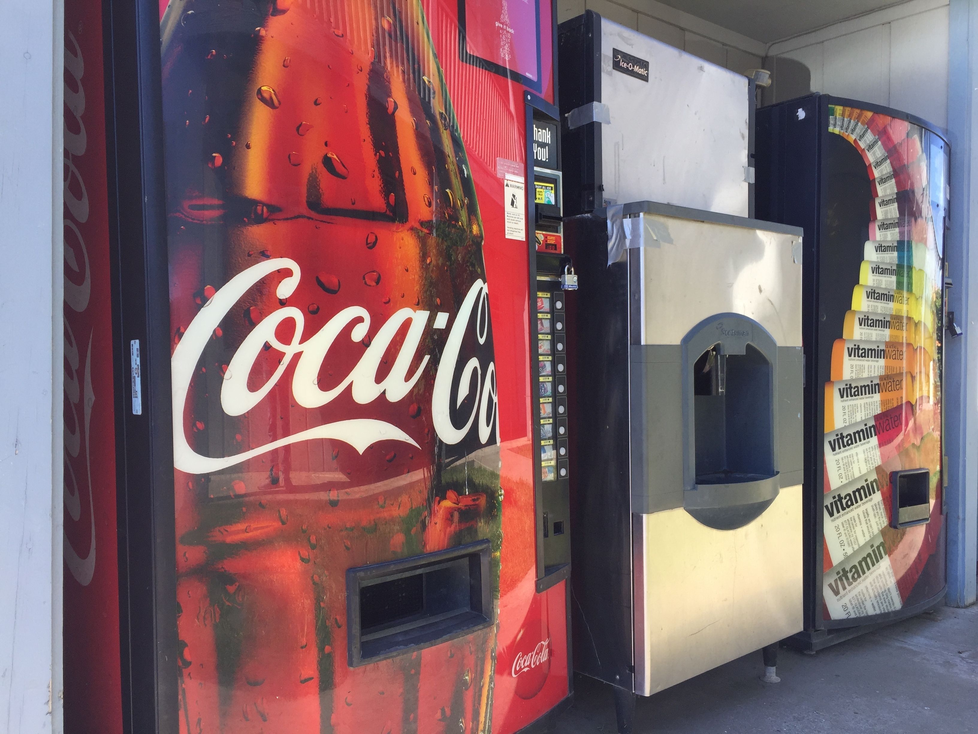 Three vending machines, one with a Coca-Cola logo, one with a vitamin water logo, and one with a generic label.