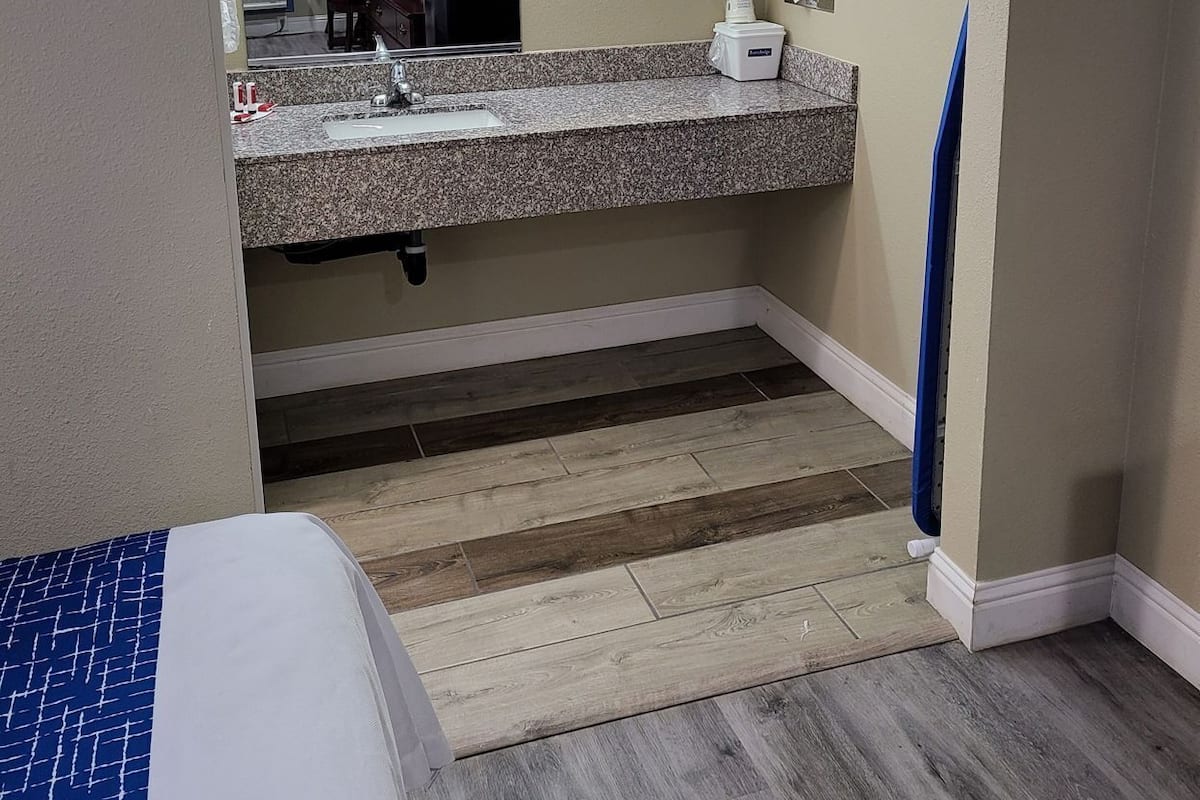 Une chambre d’hôtel avec un lavabo doté d’un plan de travail en granit, un miroir, un porte-serviettes, une petite table avec une tasse et un rideau bleu.
