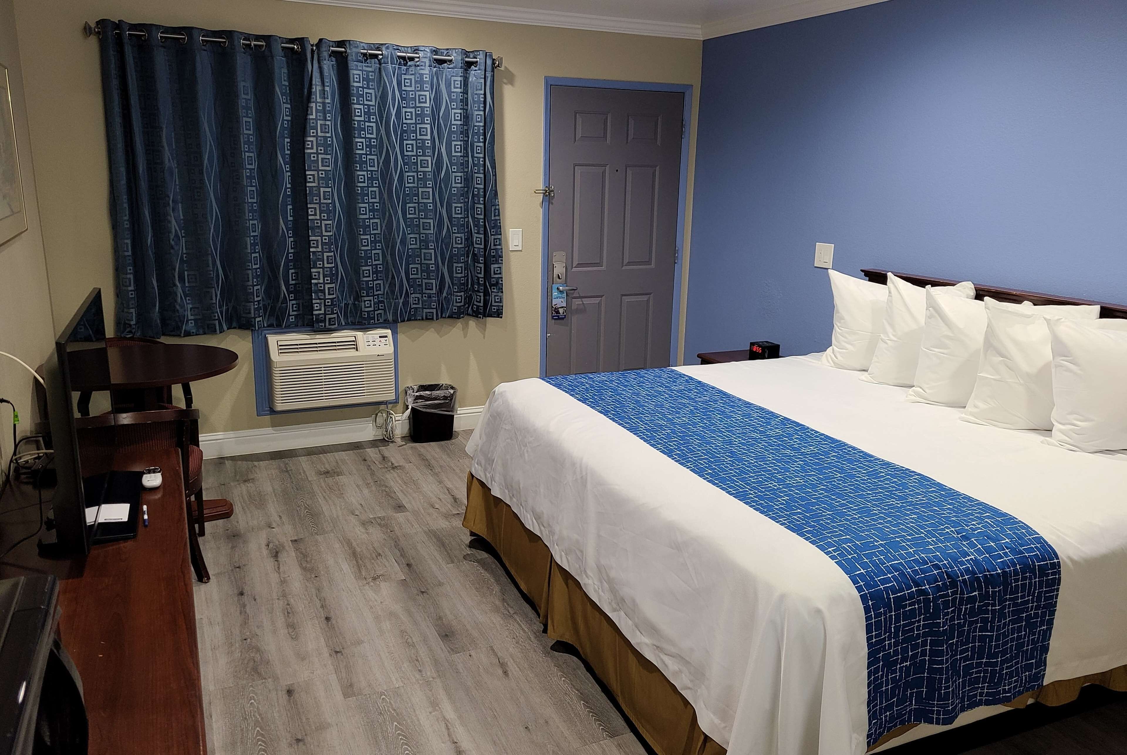 A hotel room with a bed, a desk, a chair, a window with curtains, and an air conditioning unit.