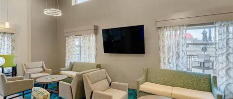 A hotel room with a large flat-screen TV, multiple seating areas including a sofa and chairs, a small table, and a patterned carpet.