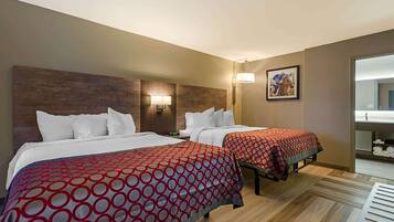A hotel room with two beds, each with white bedding and red circular-patterned bedspreads. There is a wooden headboard, a wall-mounted lamp, and a framed picture of a horse race.