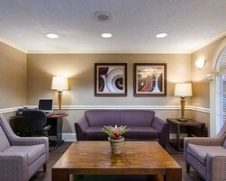 Un moderno vestíbulo de hotel con un sofá, dos sillones, una mesa baja con una planta, un escritorio con una computadora y dos cuadros abstractos en la pared.