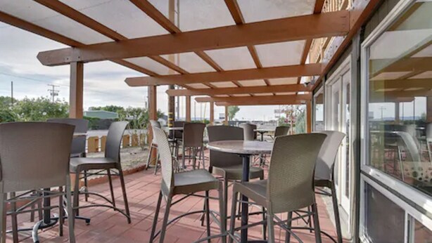 Un área de patio al aire libre con mesas y sillas de estructura metálica, una pérgola de madera y ventanas amplias que ofrecen vistas del paisaje circundante.