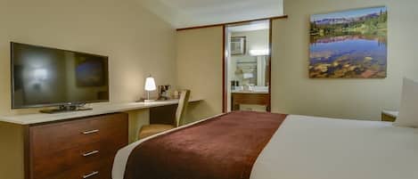 Habitación de hotel con cama, escritorio con computadora, televisor y un cuadro de paisaje.