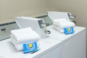 Une machine à laver à chargement frontal, avec une pile de serviettes blanches et une bouteille de lessive All.