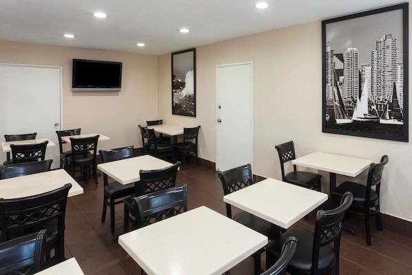A dining area with multiple tables and chairs, a flat-screen TV mounted on the wall, and a framed cityscape picture on the wall.