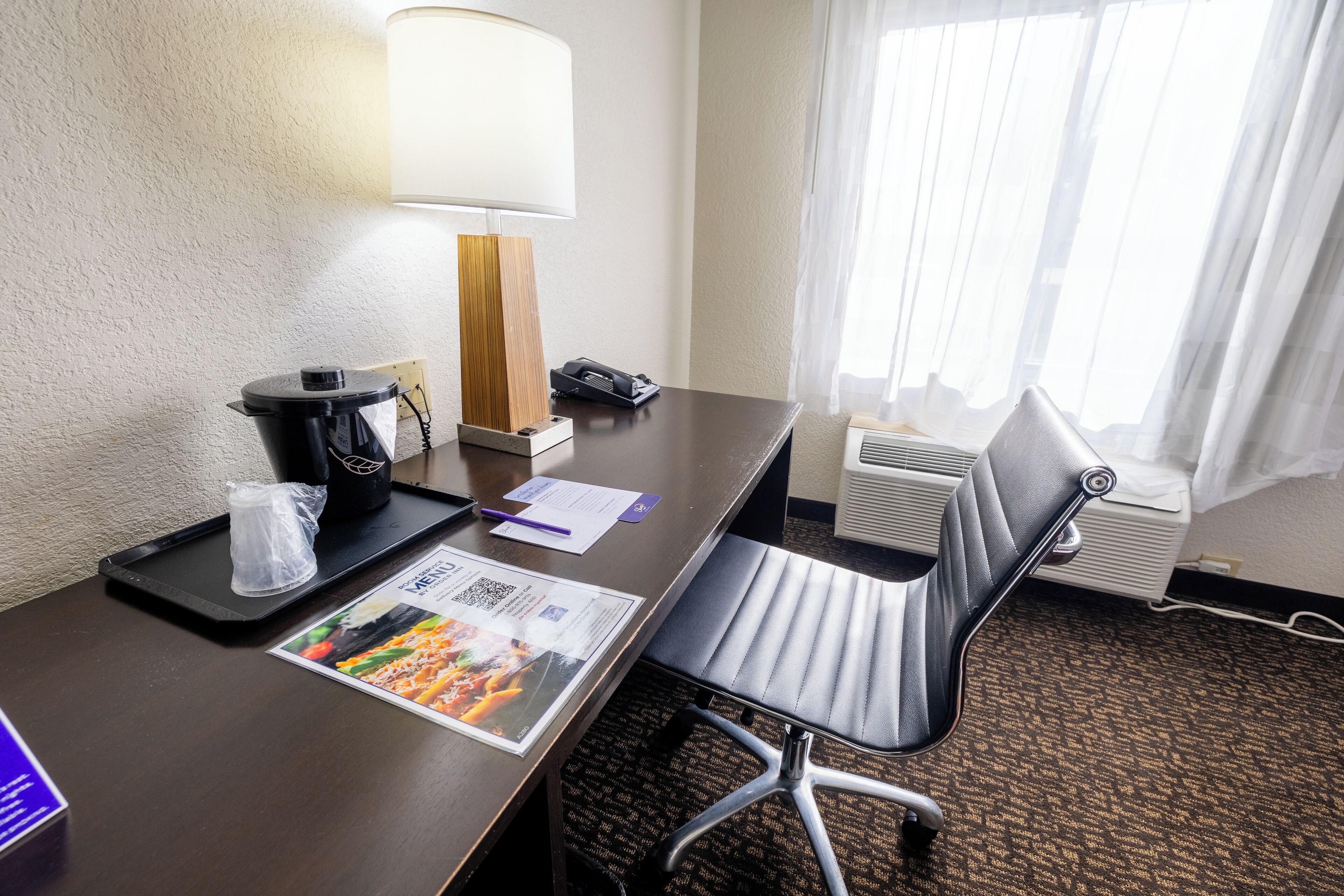Una habitación de hotel con un escritorio de madera oscura, una silla de oficina negra, una lámpara, un teléfono, una bandeja con una bolsa de plástico y un menú sobre la mesa.