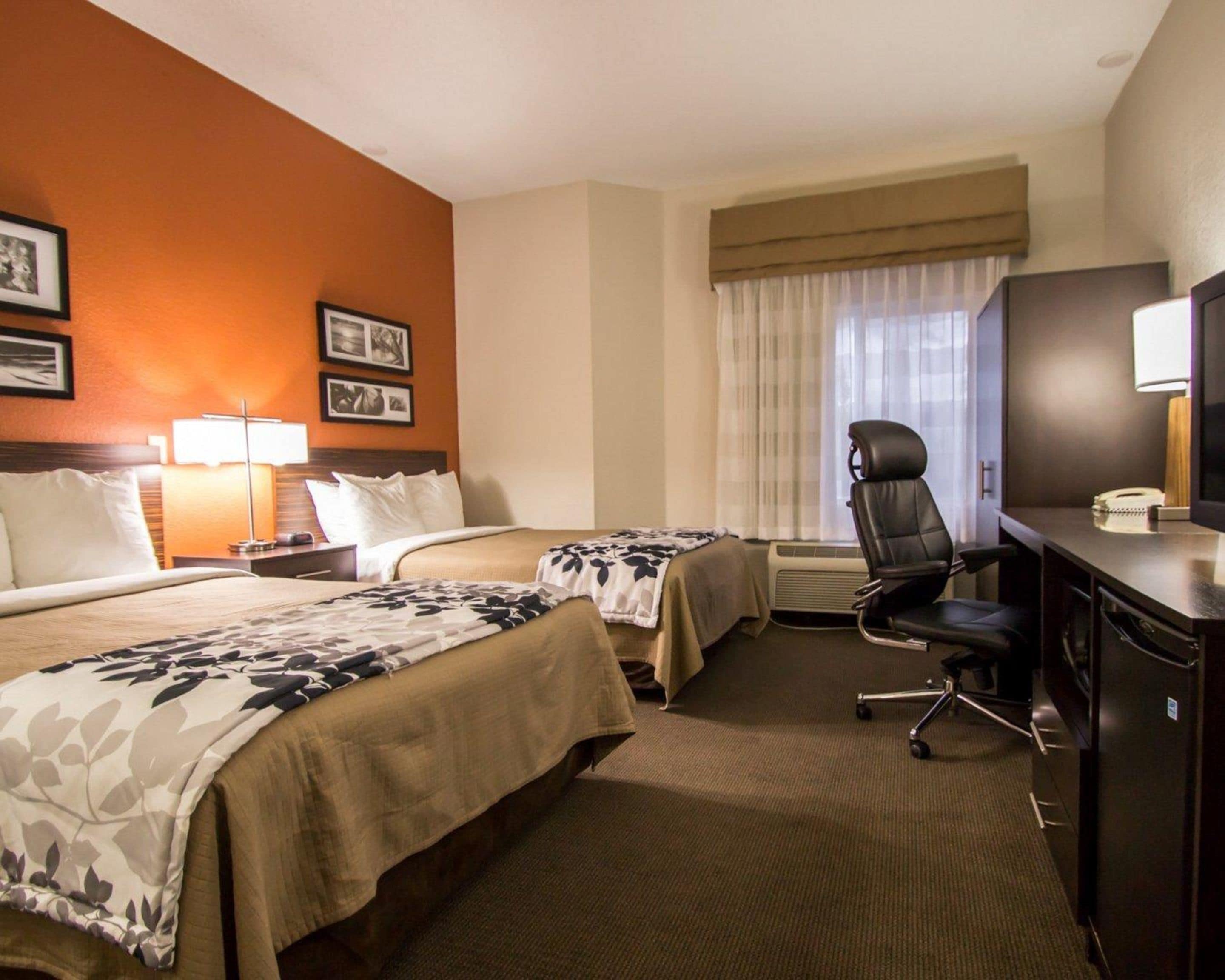 A hotel room with two beds, a desk with a chair, a desk lamp, a refrigerator, and a window with curtains.