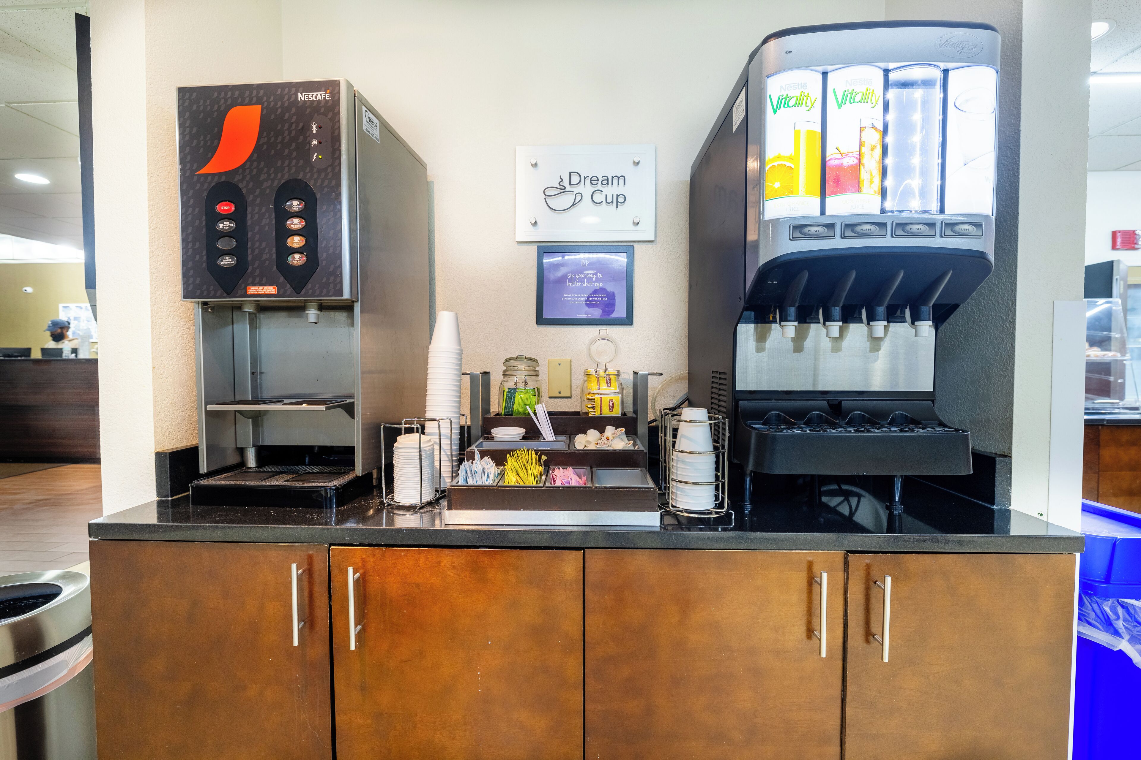 A coffee station with a Nescafé machine and a Vitality water dispenser, a Dream Cup sign, and a counter with various items.