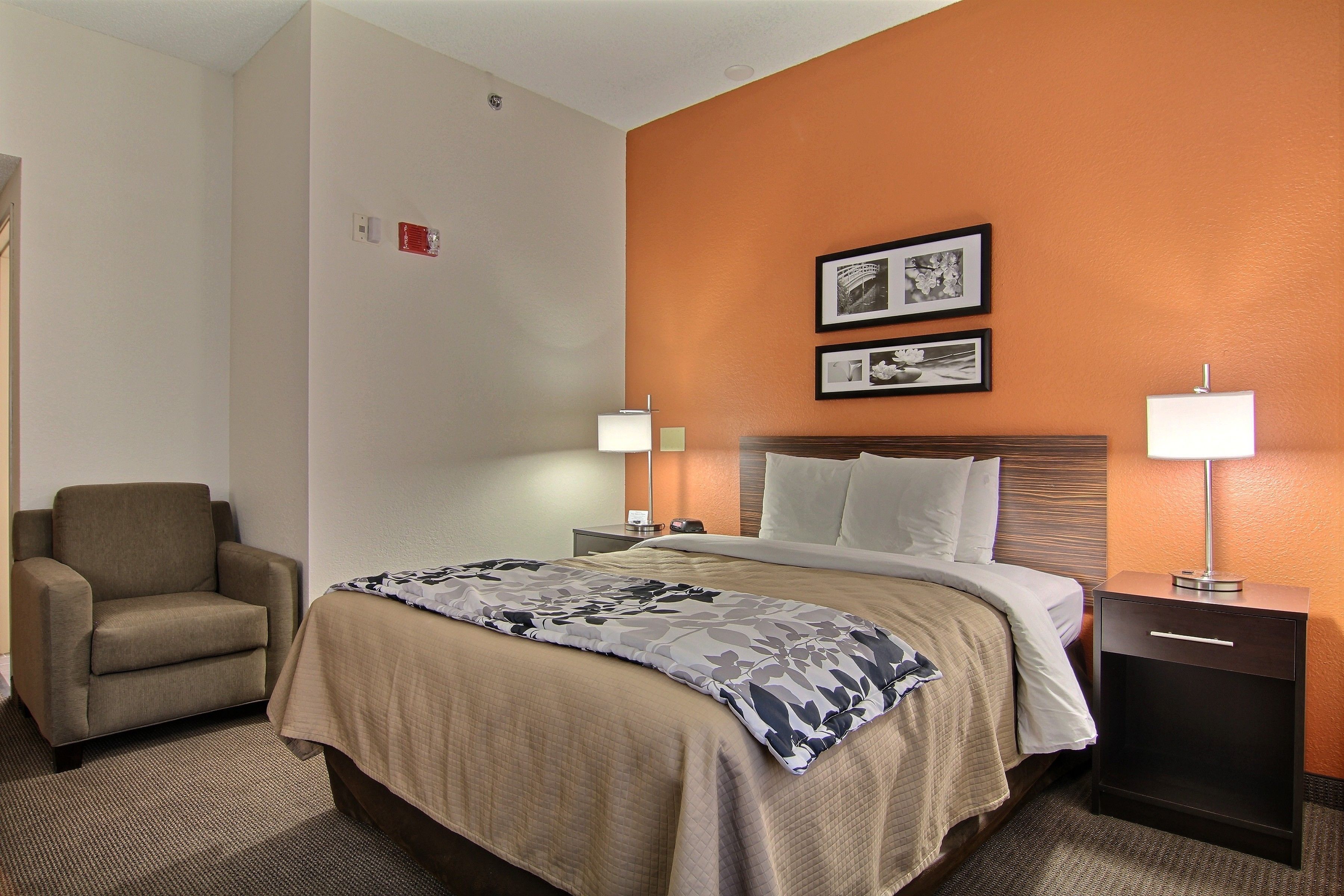 A hotel room with a bed, a chair, a nightstand with a lamp, and two framed pictures on the wall.