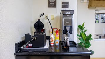 En frukostbar med våffeljärn, kaffemaskin och olika tillbehör. Det finns skyltar som anger tidpunkterna för den kostnadsfria frukosten och det maximala antalet personer som får vistas där samtidigt.