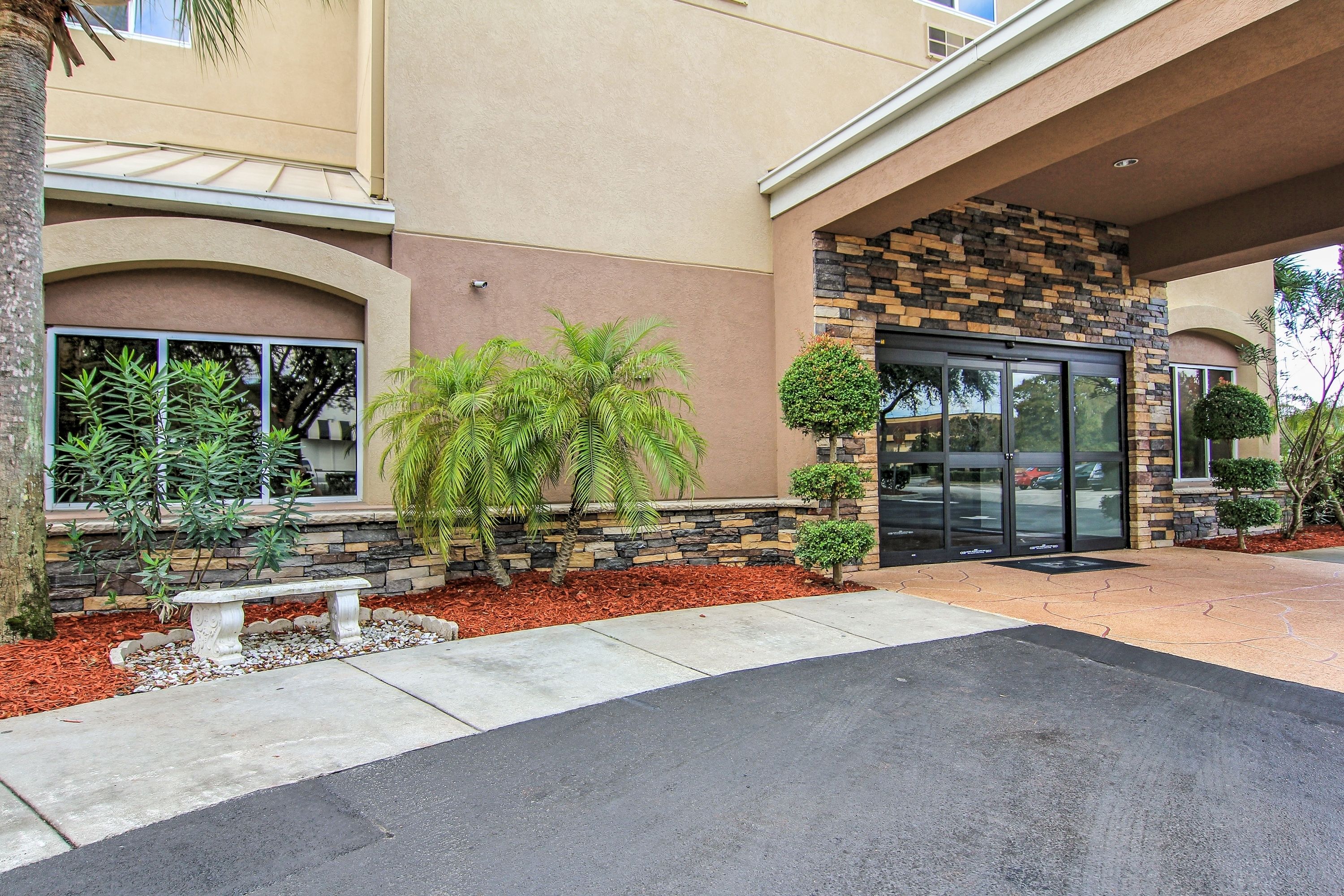 A building entrance with a stone facade, a palm tree, and a bench.