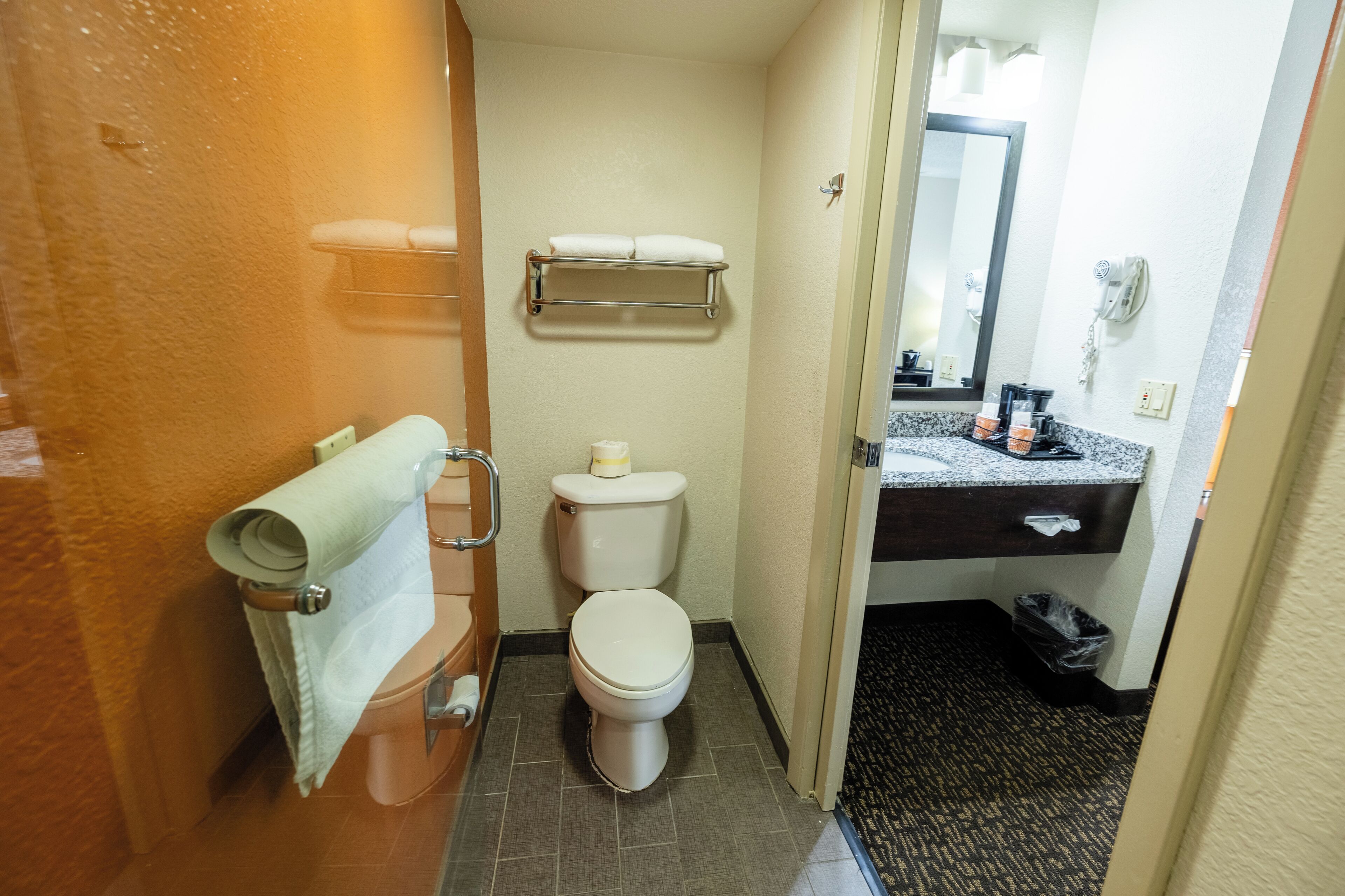 A compact bathroom with a toilet, a towel rack, a small shelf with towels, a mirror, and a hairdryer.