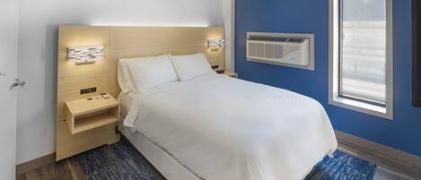 A modern hotel room with a large bed, bedside tables, a nightstand, a wall-mounted air conditioner, and a window with blinds.