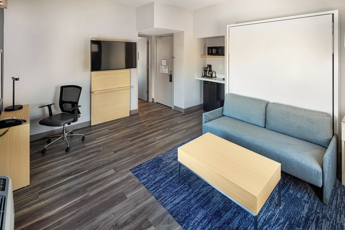A modern living room with a blue sofa, a wooden coffee table, a black office chair, a flat-screen TV, and a kitchenette with a microwave.