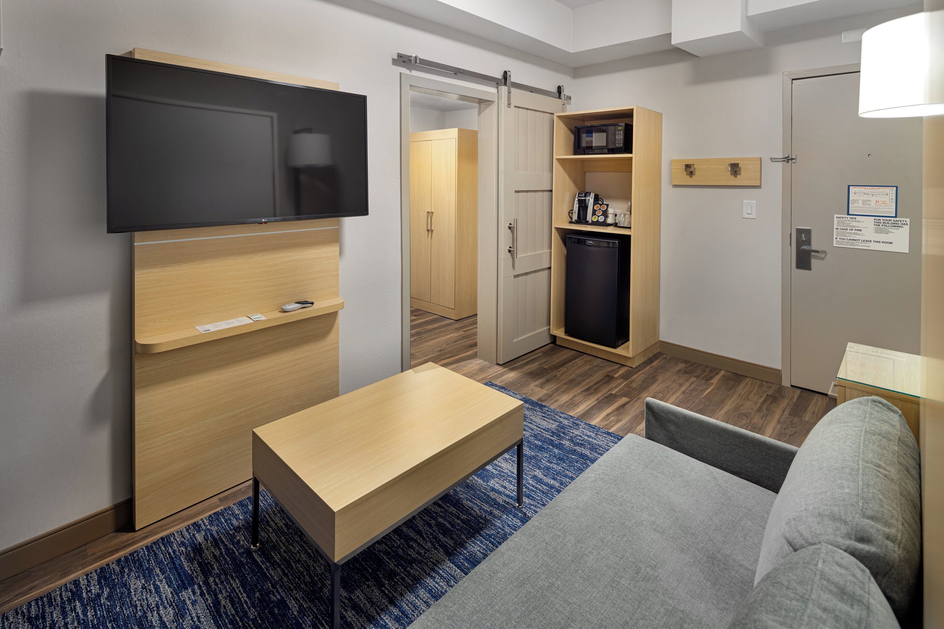 A modern hotel room with a flat-screen TV, a small wooden table, a sofa, a kitchenette with a microwave, and a sliding door closet.