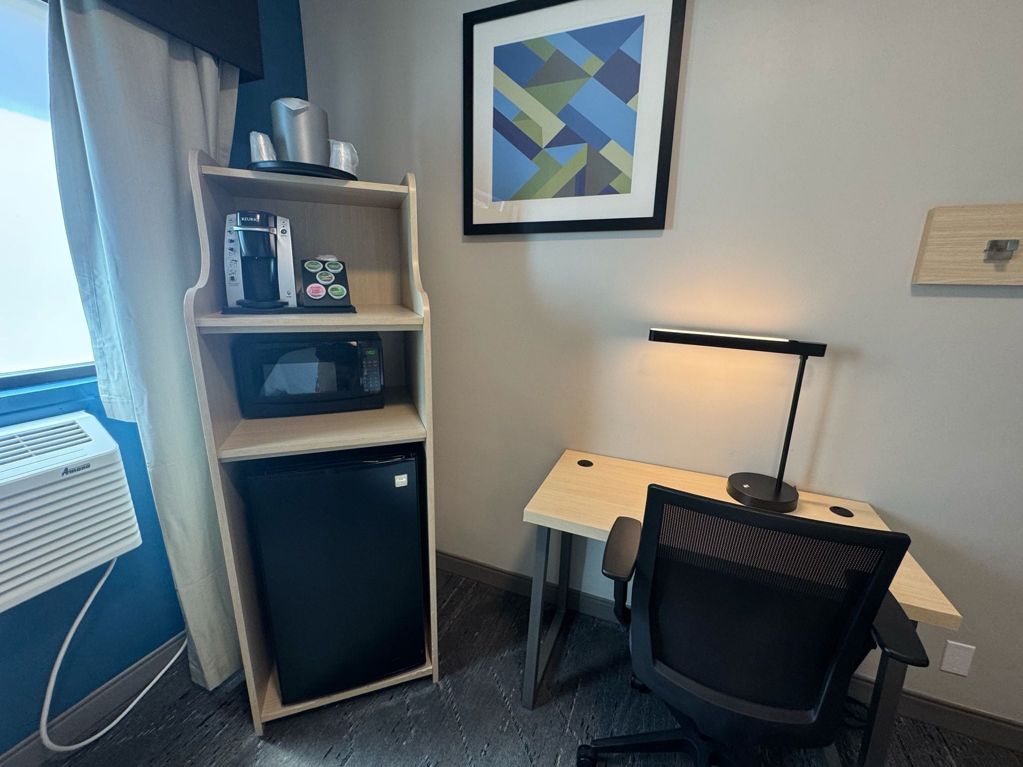 A compact hotel room with a kitchenette, a desk with a chair, a framed abstract painting, and a standing lamp.