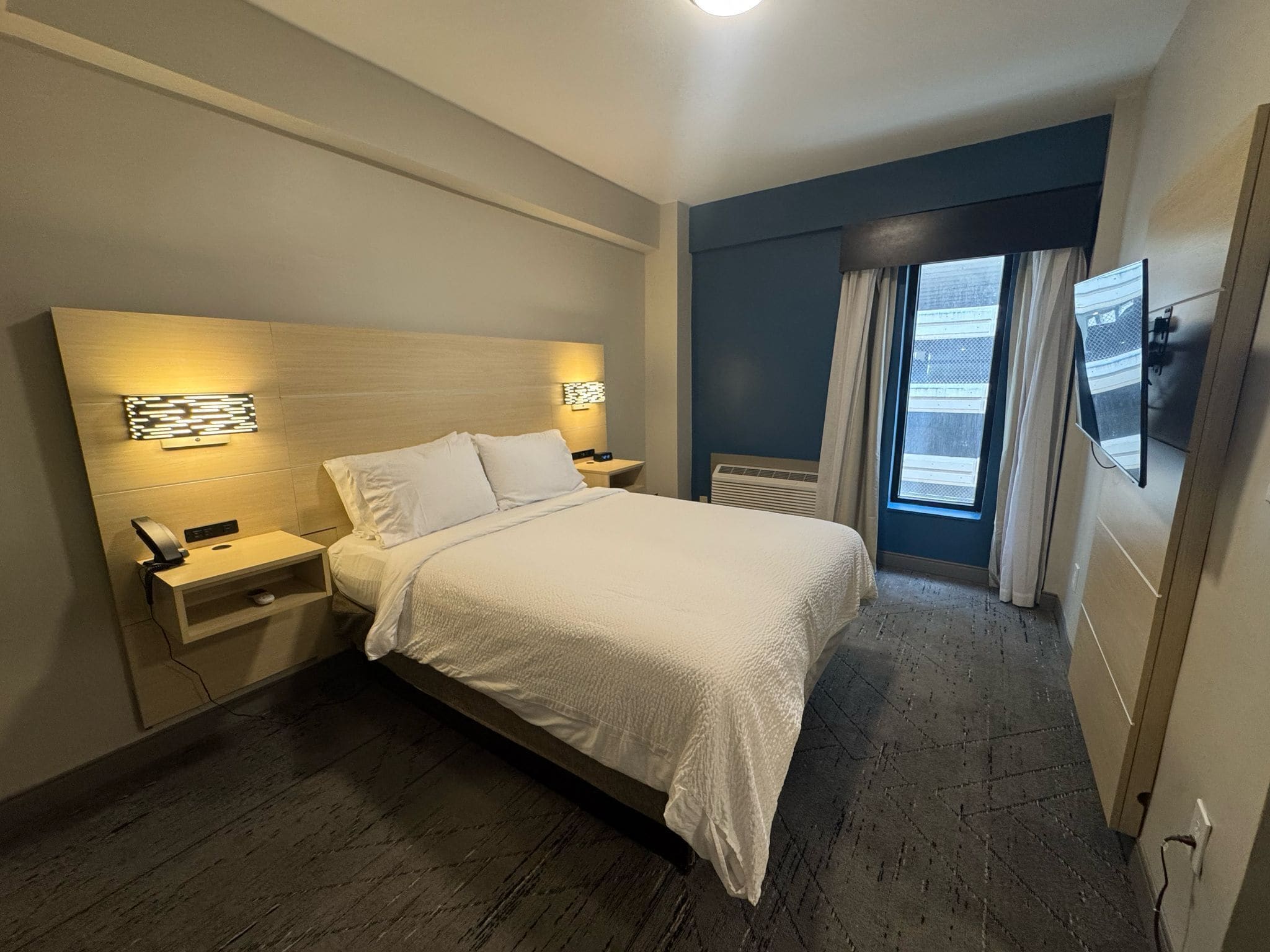 A hotel room with a bed, bedside tables, a wall-mounted light, a window with curtains, and a view of the outside.