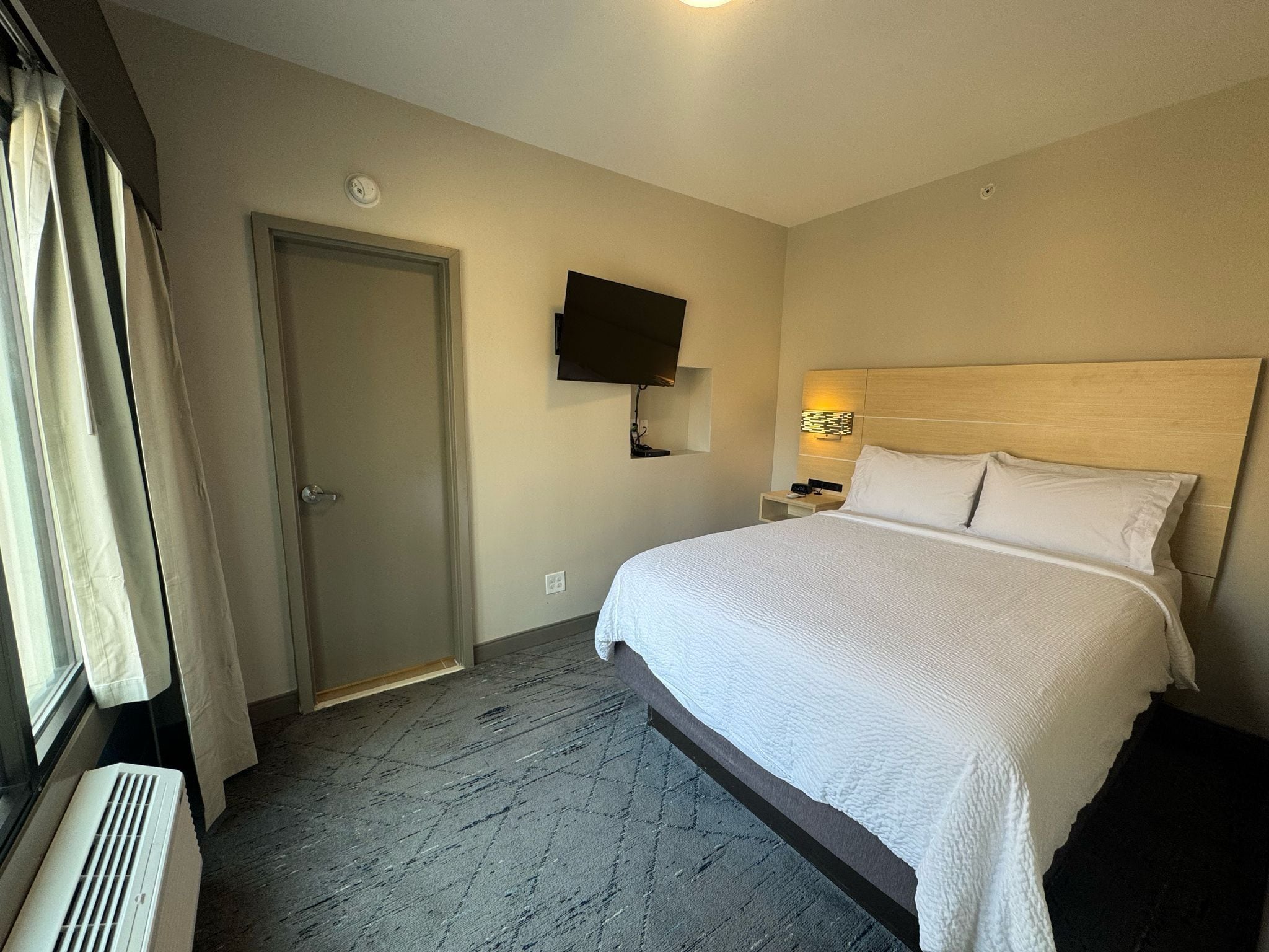 A hotel room with a large bed, a flat-screen TV mounted on the wall, a bedside table with a lamp, and a door.