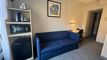 A hotel room with a blue sofa, a kitchenette with a microwave, a coffee maker, and a small refrigerator. There is a framed abstract artwork on the wall and a desk with a lamp.