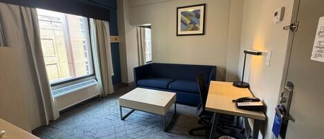 Une chambre d’hôtel avec un canapé bleu, une petite table, un lampadaire et une fenêtre avec des rideaux.