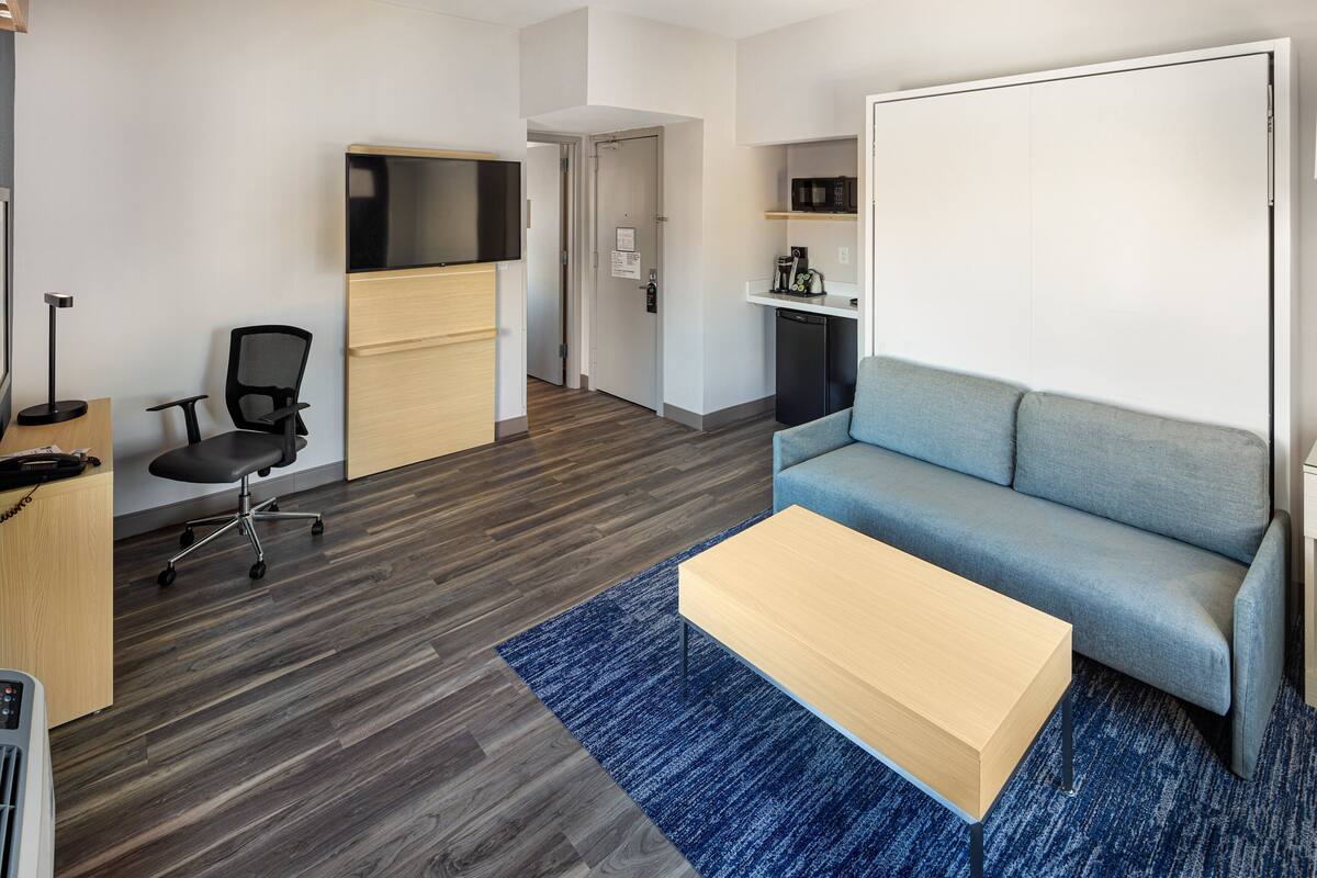 A modern living room with a blue sofa, a wooden coffee table, a black office chair, a flat-screen TV, and a kitchenette with a microwave.