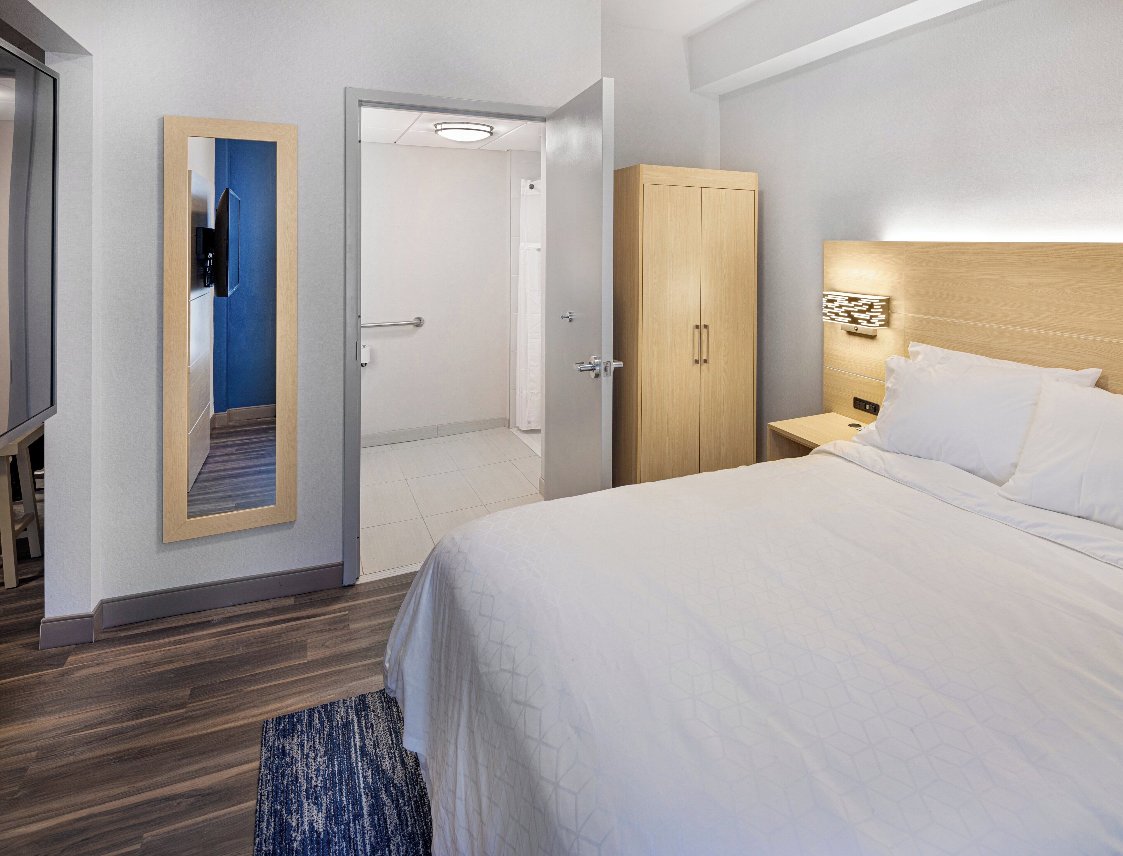 A hotel room with a bed, a wardrobe, a nightstand, a mirror, and a door leading to a bathroom.